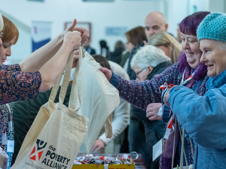 Poverty Alliance staff helping delegates register at our 2022 conference. A young woman handing over a Poverty Alliance tote bag to an older woman dressed all in blue, with a blue hat.